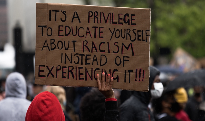 A Black person is seen from behind at a protest, holding a sign. It reads: it's a privilege to educate yourself about racism instead of experiencing it