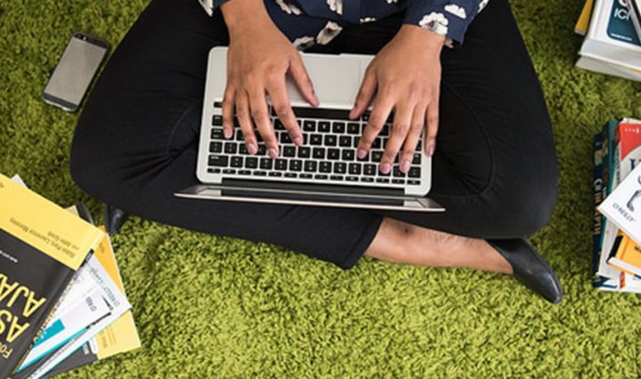 Black woman's hands are typing on a laptop. She sits on the ground surrounded by books