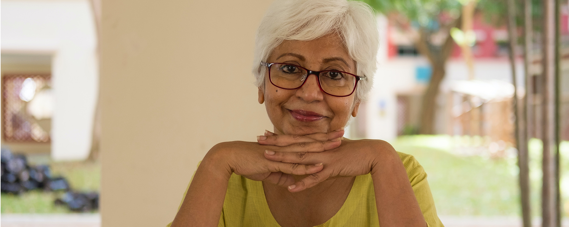 Older South Asian woman with dark skin wears glasses and smiles with her hands folded