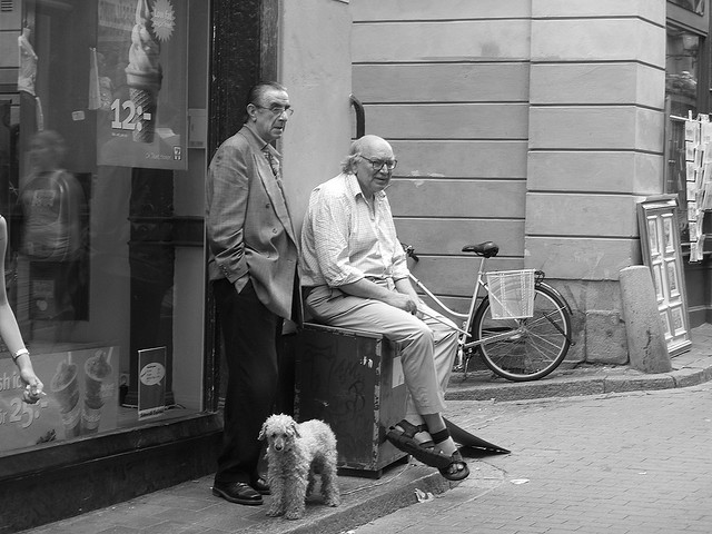 Two elderly men from Sweden are sitting in front of a shop with a small dog at their feet