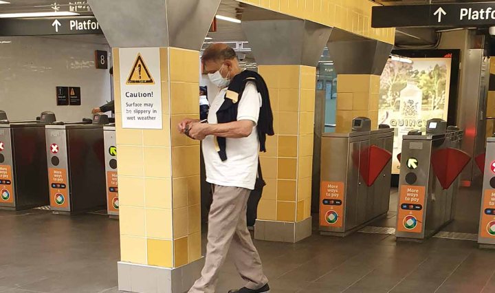 A Brown man walks across Central Station wearing a face mask. A sign behind says "Caution"